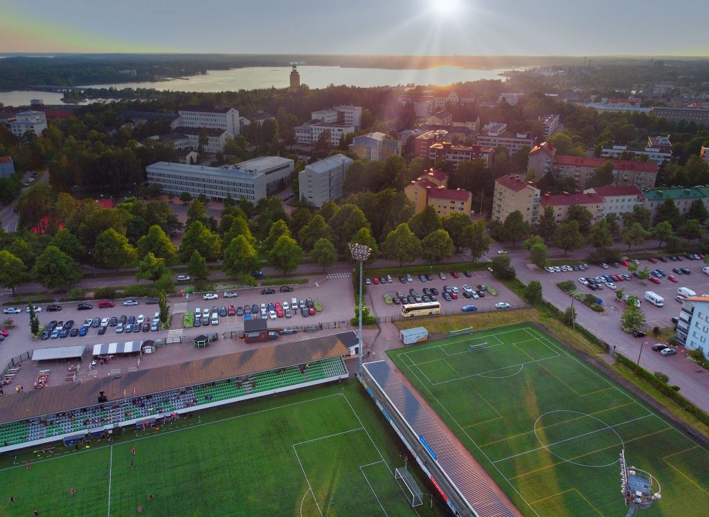 Jalkapallokenttä kuvattuna ylhäältä päin. Taustalla auringonlasku ja Kotkan kaupunki.
