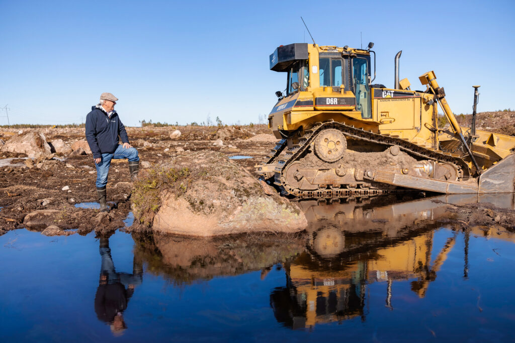 Ihminen maanrakennuskoneen vieressä. Etualalla vettä.