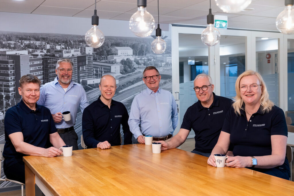 Ryhmä työntekijöitä istumassa pöydän ääressä ja katsomassa kameraan.